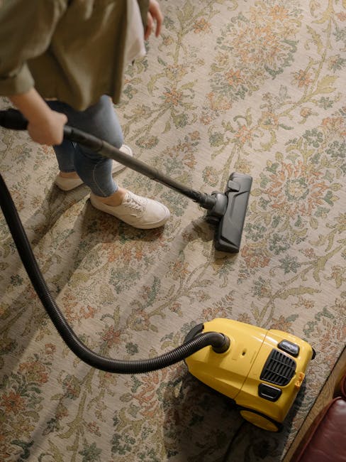 A person wearing beige shoes and blue jeans is using a vacuum cleaner on a traditional patterned area rug with cream, green, and orange tones in a room. The vacuum cleaner, yellow with black accents, 