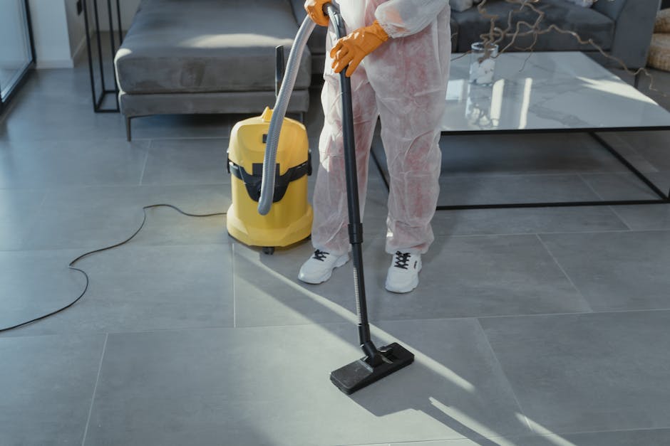 A person wearing beige shoes and blue jeans is using a vacuum cleaner on a traditional patterned area rug with cream, green, and orange tones in a room. The vacuum cleaner, yellow with black accents, 