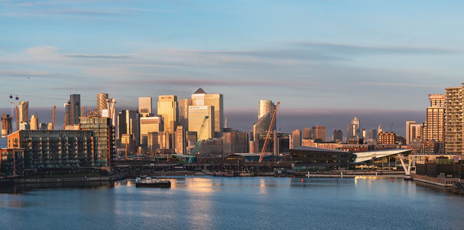 A panoramic view of London's financial district with modern skyscrapers, including the Walkie Talkie building and the Leadenhall Building, taken from across the River Thames during sunset. The water r