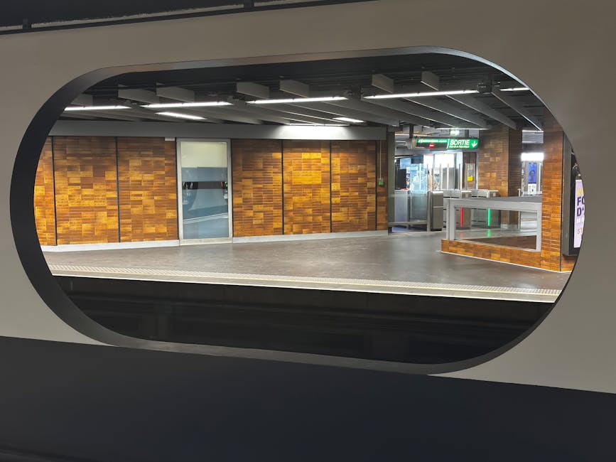 View of Prince Regent Station platform seen through an oval window, featuring wooden-paneled walls with a polished finish, a smooth grey concrete floor, illuminated ceiling panels, and an emergency ex
