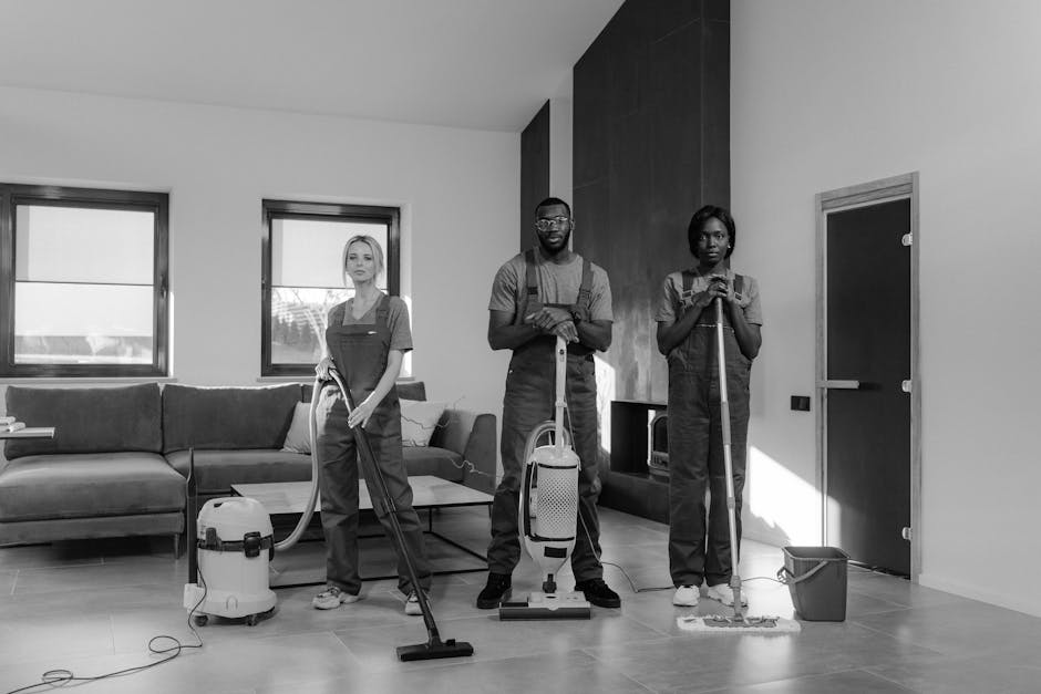 A person, wearing beige shoes and casual clothing, is using a yellow vacuum cleaner with a black hose to clean a traditional patterned carpet in a residential living room. The carpet features a detail